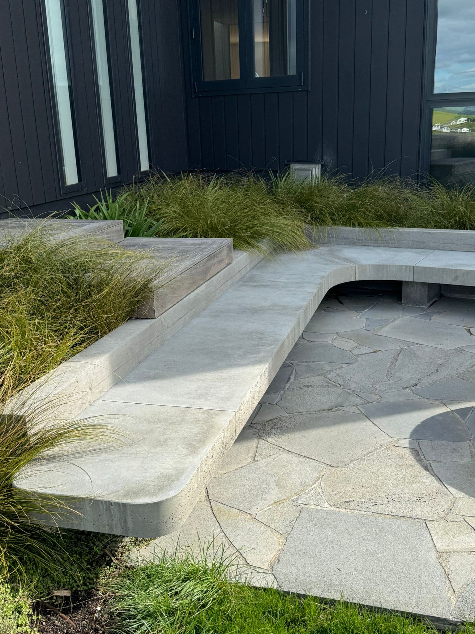 Concrete bench seating with bluestone paving and native grasses alongside modern dark-clad Auckland home